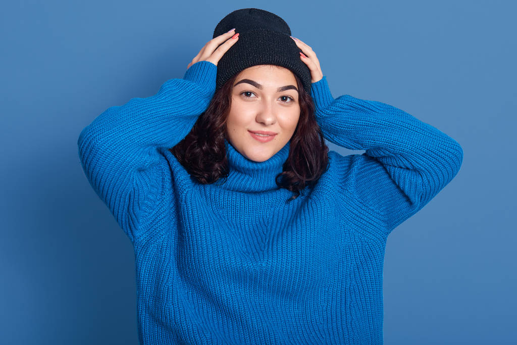 Poziomy obraz pięknej kobiety noszącej ciemnoniebieski sweter, patrzącej bezpośrednio w aparat, trzymającej ręce na głowie, o kręconych czarnych włosach, będącej w dobrym nastroju. Koncepcja zimowa. - Zdjęcie, obraz