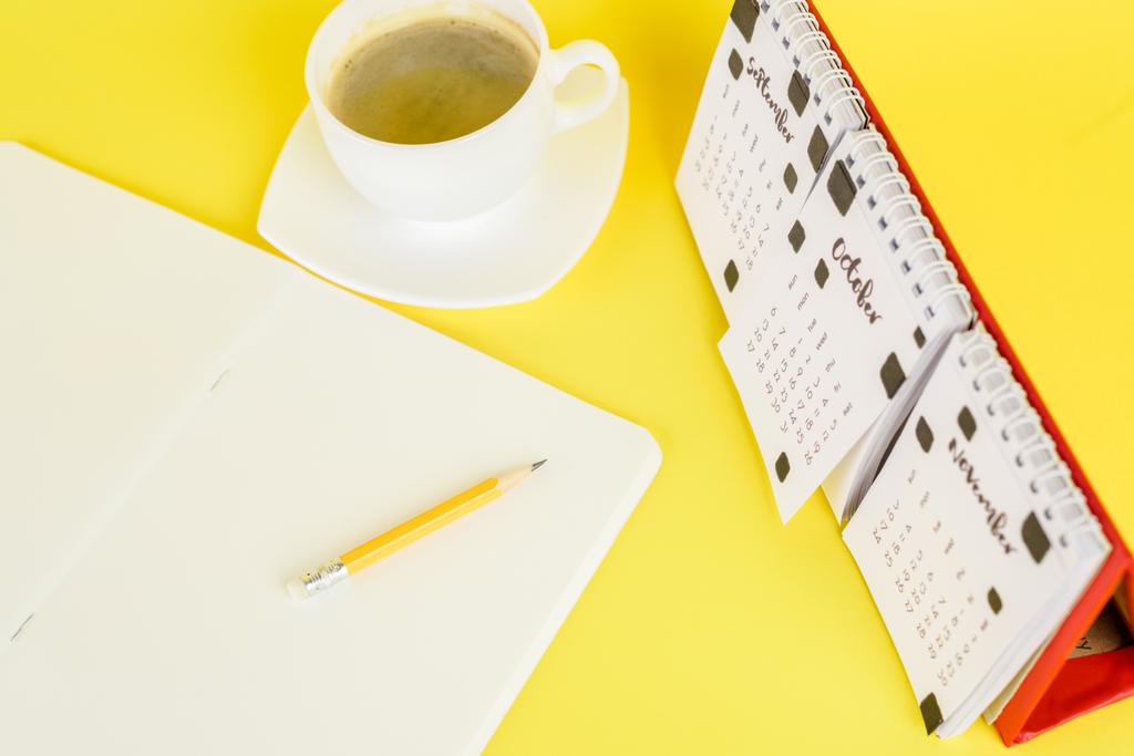Blick auf leeres Notizbuch in der Nähe von Kaffeetasse und Kalender auf gelbem Hintergrund - Foto, Bild
