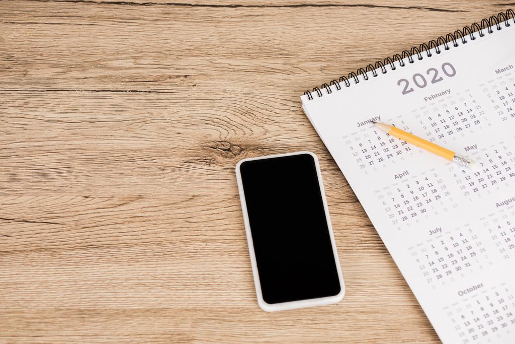 Υψηλής γωνίας άποψη του smartphone με κενή οθόνη και το ημερολόγιο του 2020 έτος με μολύβι σε ξύλινο φόντο - Φωτογραφία, εικόνα