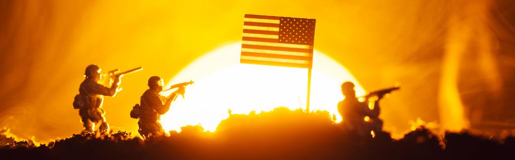 Scena bitwy z zabawkowymi wojownikami w pobliżu amerykańskiej flagi z dymem i zachodem słońca na tle, panoramiczne ujęcie - Zdjęcie, obraz
