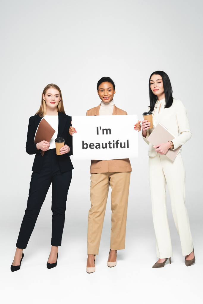 fröhliche multikulturelle Geschäftsfrauen mit Ordnern und Pappbechern in der Nähe von Plakaten mit dem schönen Schriftzug "I 'm beautiful" auf weißem Grund  - Foto, Bild
