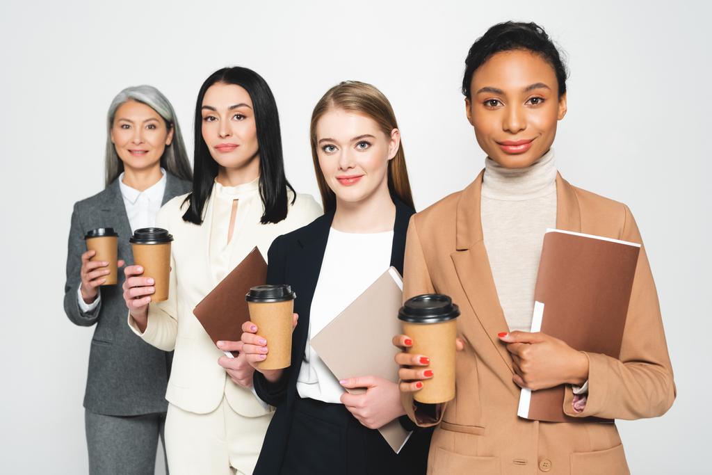 four happy and multicultural businesswomen holding folders and paper cups isolated on white  - Photo, Image
