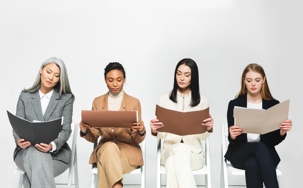 four multicultural businesswomen in suits sitting on chairs and looking at folders isolated on white  - Photo, Image