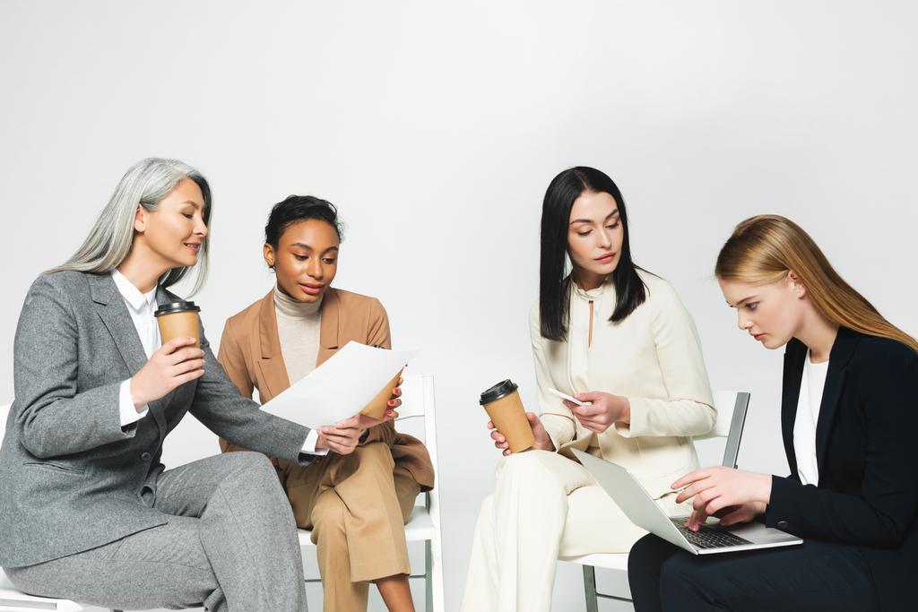multiculturele zakenvrouwen zitten op stoelen met papieren bekers en laptop geïsoleerd op wit  - Foto, afbeelding