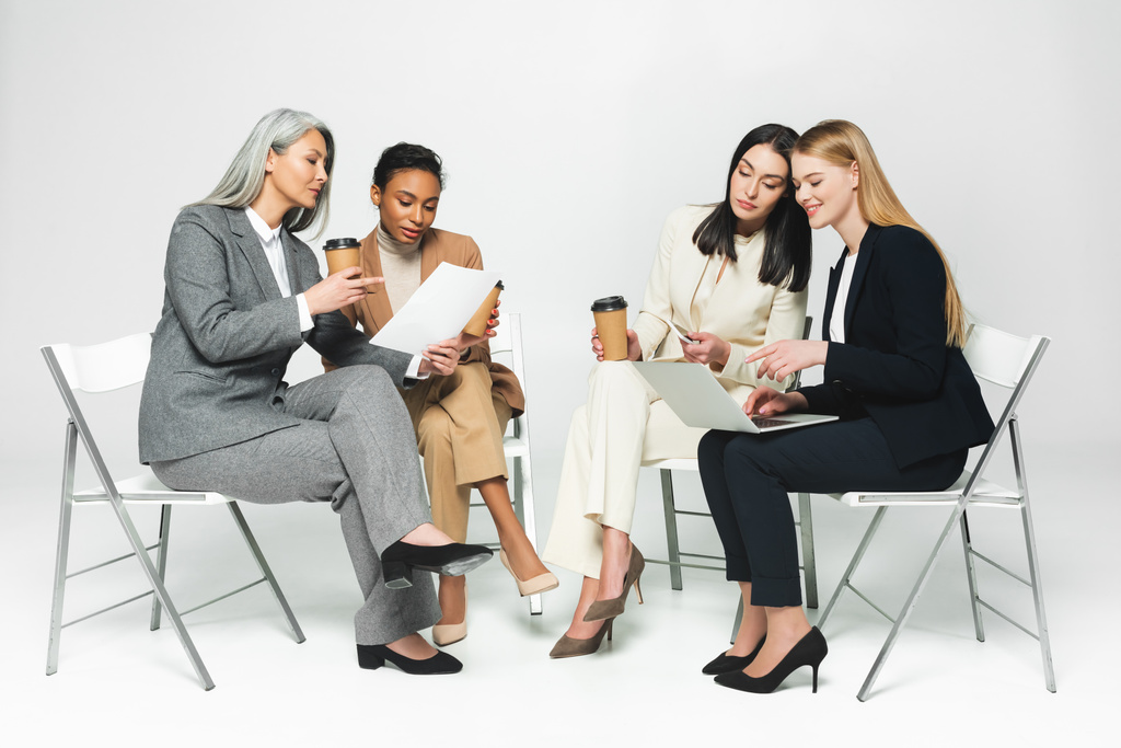 mulheres de negócios multiculturais felizes sentadas em cadeiras com copos de papel e laptop em branco
  - Foto, Imagem