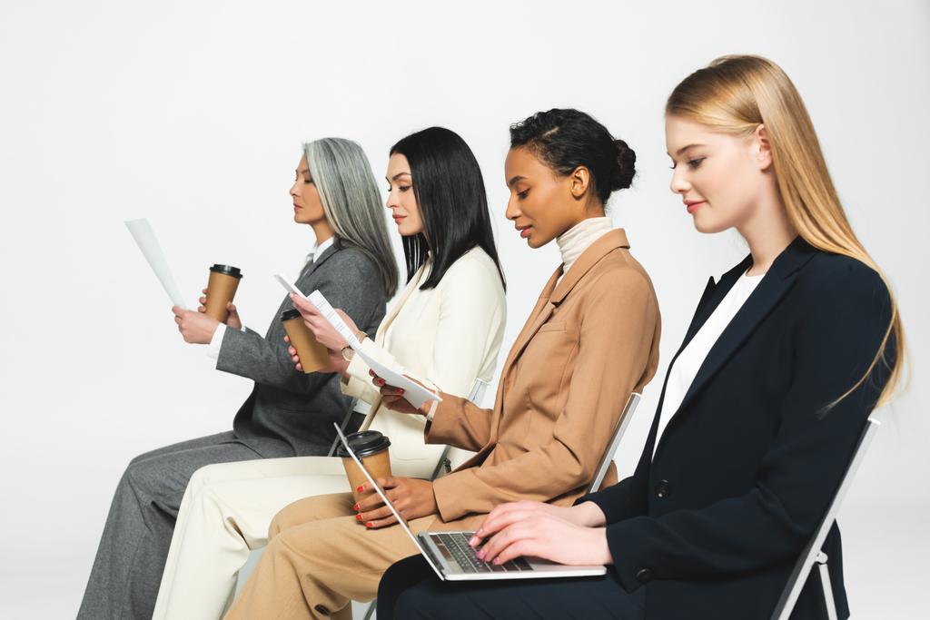 вид збоку мультикультурних бізнесменів, що сидять на стільцях з паперовими чашками та ноутбуком на білому
  - Фото, зображення