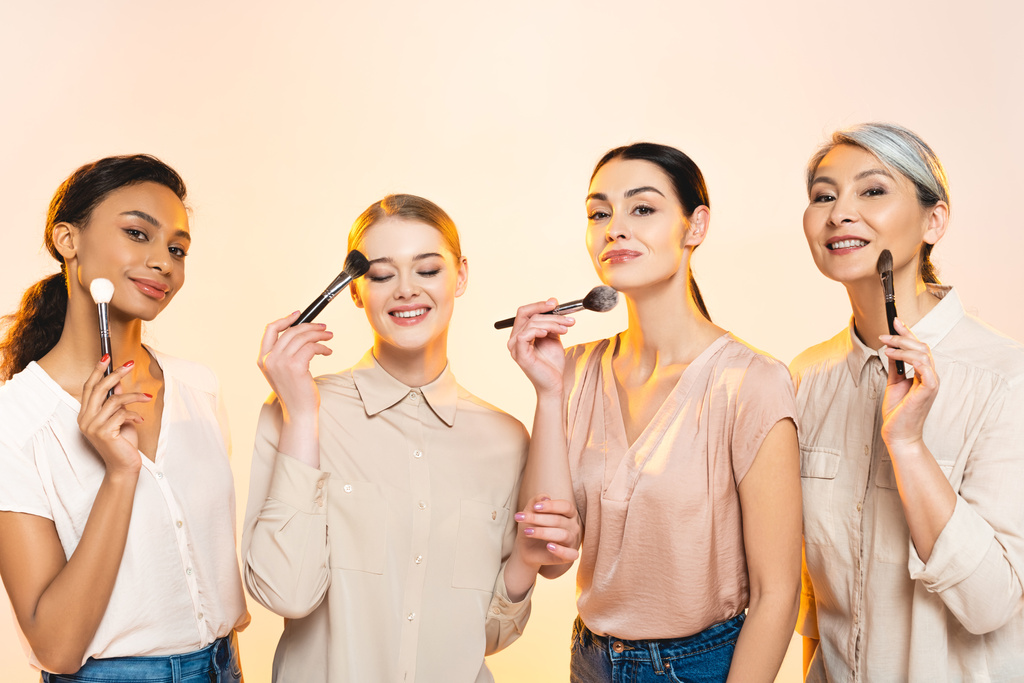 glückliche multikulturelle Frauen mit Kosmetikbürsten isoliert auf Beige  - Foto, Bild