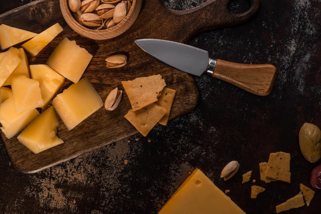 Bıçağın yanındaki tahta kesme tahtasının üzerinde fıstıklı ve krakerli peynir tabağının üst görüntüsü. - Fotoğraf, Görsel