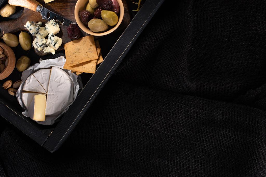 Top view of tray with pieces of camembert, dorblu, dried olives, crackers and pistachios isolated on black - Photo, Image