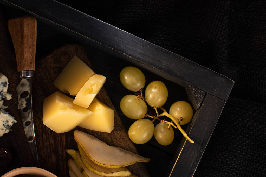 Вид зверху лотка з брудним ножем біля шматків сиру, скибочки груші і винограду ізольовані на чорному
  - Фото, зображення