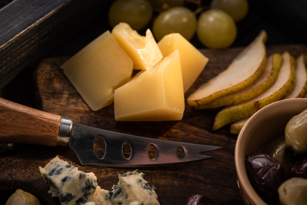 Foco seletivo de faca, pedaços de queijo, fatias de pêra, azeitonas secas, uvas na tábua de corte na bandeja
 - Foto, Imagem