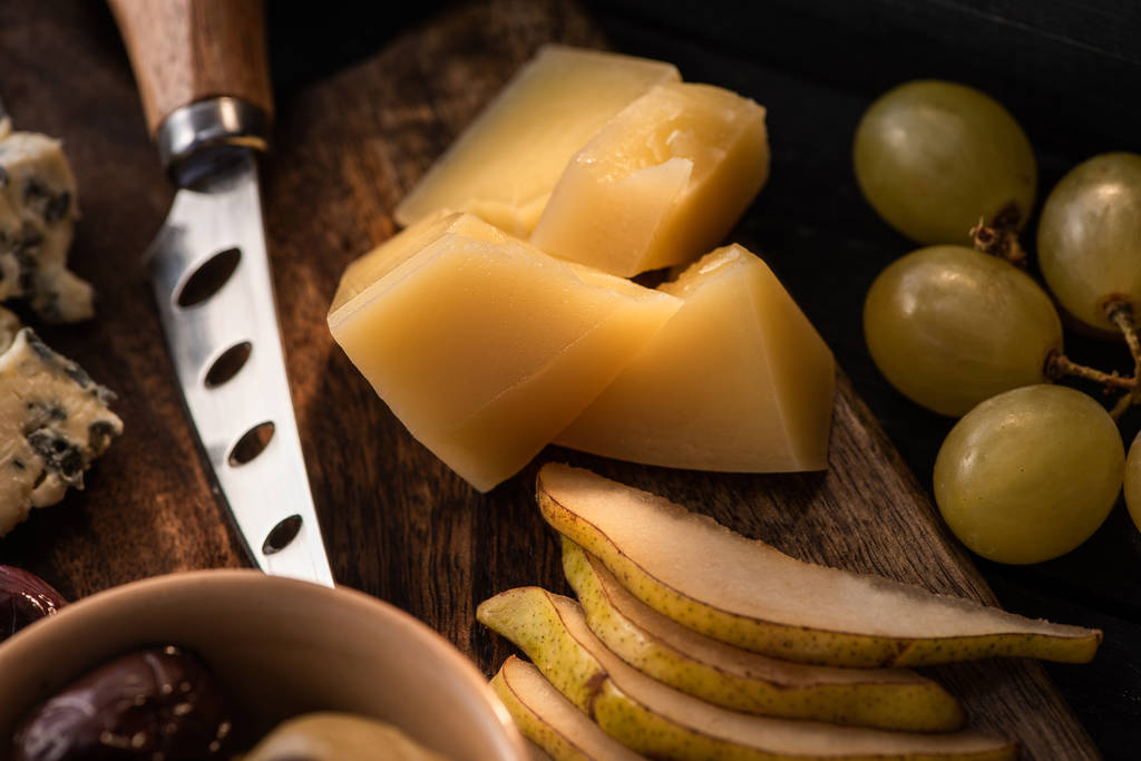 Enfoque selectivo de grana padano con trozos de pera, tazón de aceitunas, cuchillo, dorblu, uvas en tabla de cortar
  - Foto, imagen