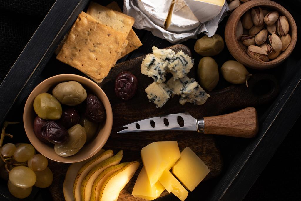 Blick von oben auf die Nahrungsmittelzusammensetzung aus Käse, getrockneten Oliven, Früchten, Pistazien und Crackern auf Holztablett  - Foto, Bild