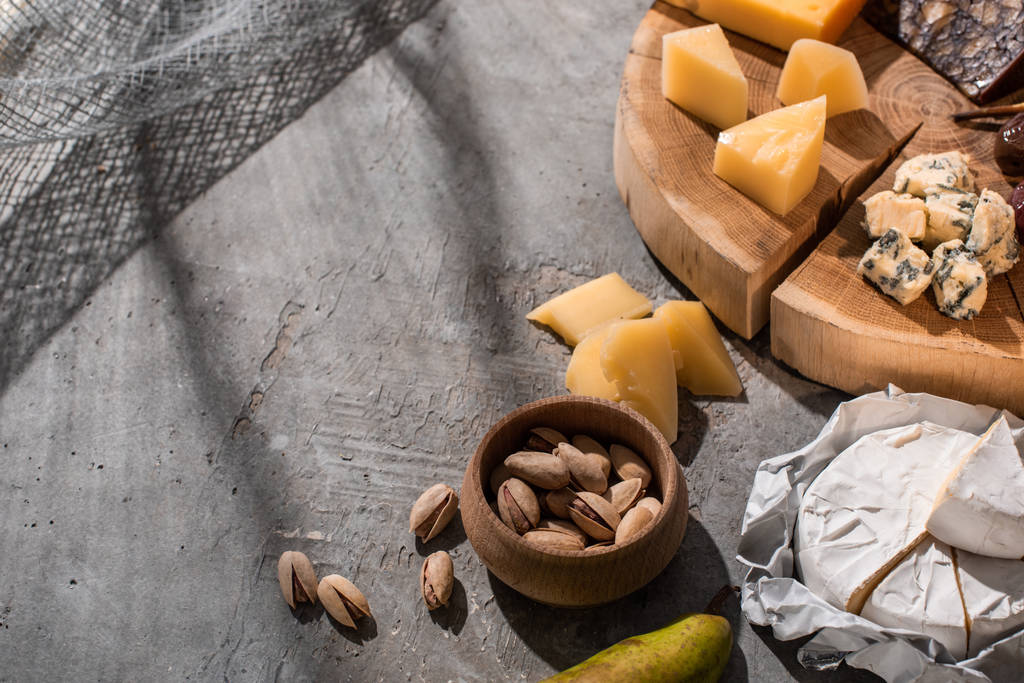 Vysoký úhel pohledu na hrušky, pistácie a camembert vedle různých druhů sýrů na dřevěné desce na šedém pozadí - Fotografie, Obrázek