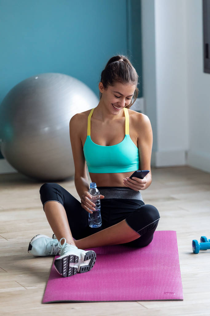 Giovane donna sportiva che usa il suo telefono cellulare mentre tiene in mano la bottiglia d'acqua dopo una lezione di pilates in palestra
. - Foto, immagini