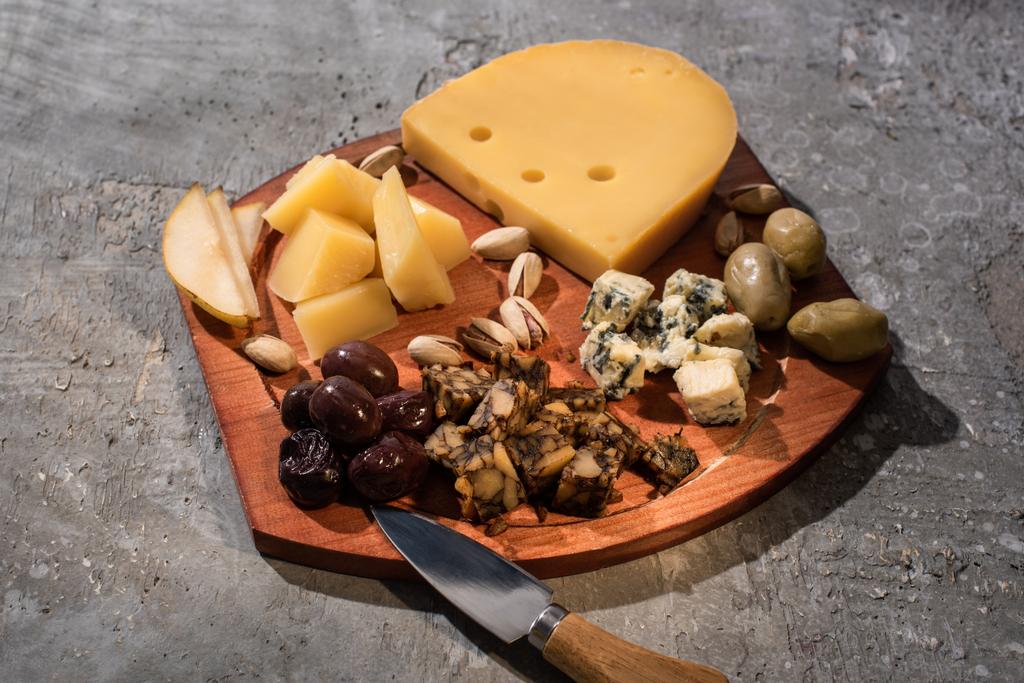 Пищевой состав сыра с сушеными оливками, кусочками груши, фисташками и ножом на разделочной доске на сером фоне
 - Фото, изображение