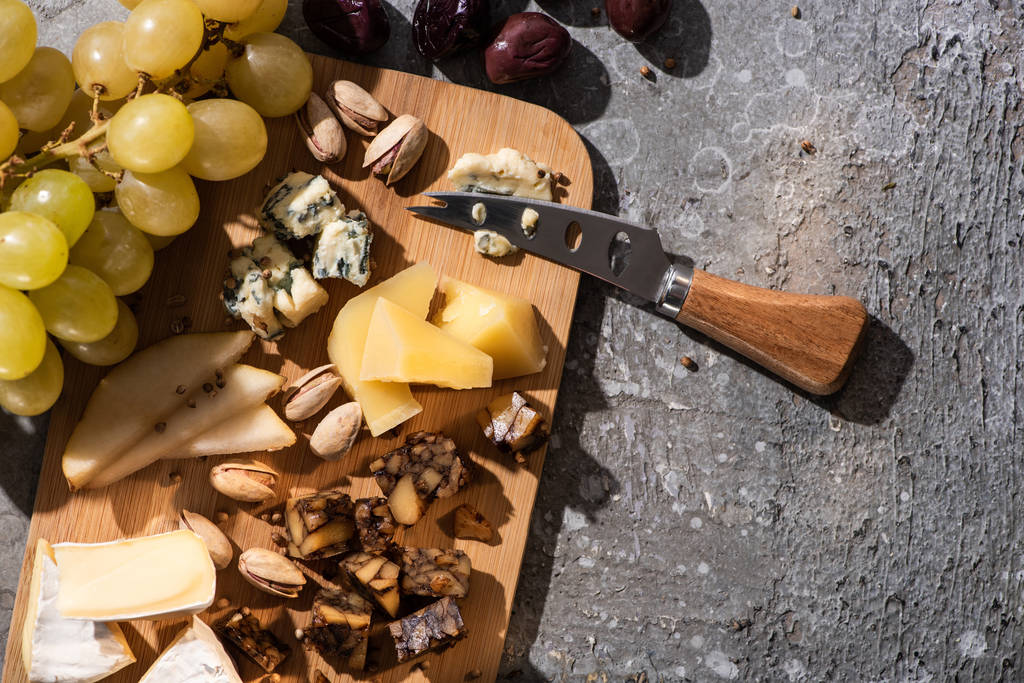 Horní pohled na kousky sýra, olivy, hrozny, pistácie, plátky hrušky a nůž na řezací desce na šedém pozadí - Fotografie, Obrázek