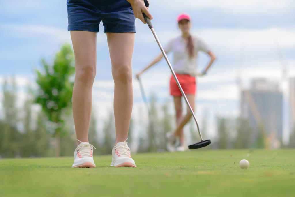 junge Golfspielerin wirft Golfball in das Loch auf dem Grün des Golfplatzes, ein Gegner oder Konkurrent wartet im Hintergrund - Foto, Bild