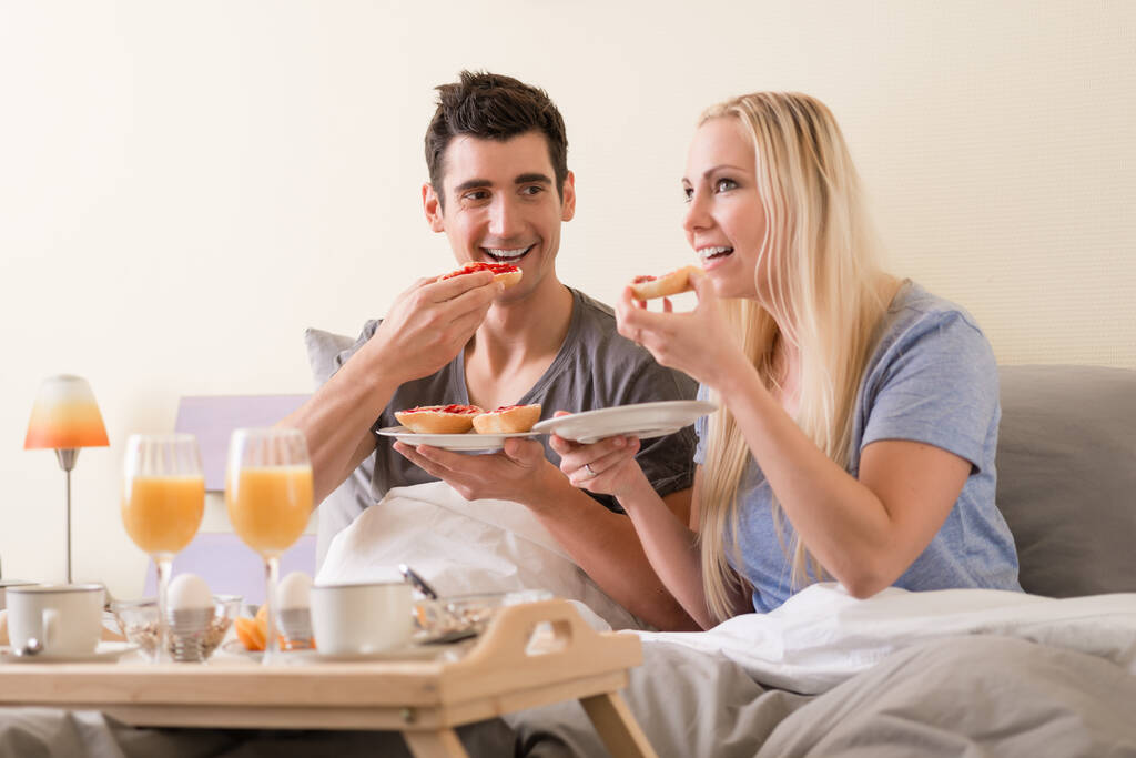 Νεαρό ζευγάρι γιορτάζει με πρωινό στο κρεβάτι κάθεται τρώει μαρμελάδα μούρο o τοστ συνοδεύεται από φρέσκο χυμό πορτοκαλιού και βραστά αυγά - Φωτογραφία, εικόνα