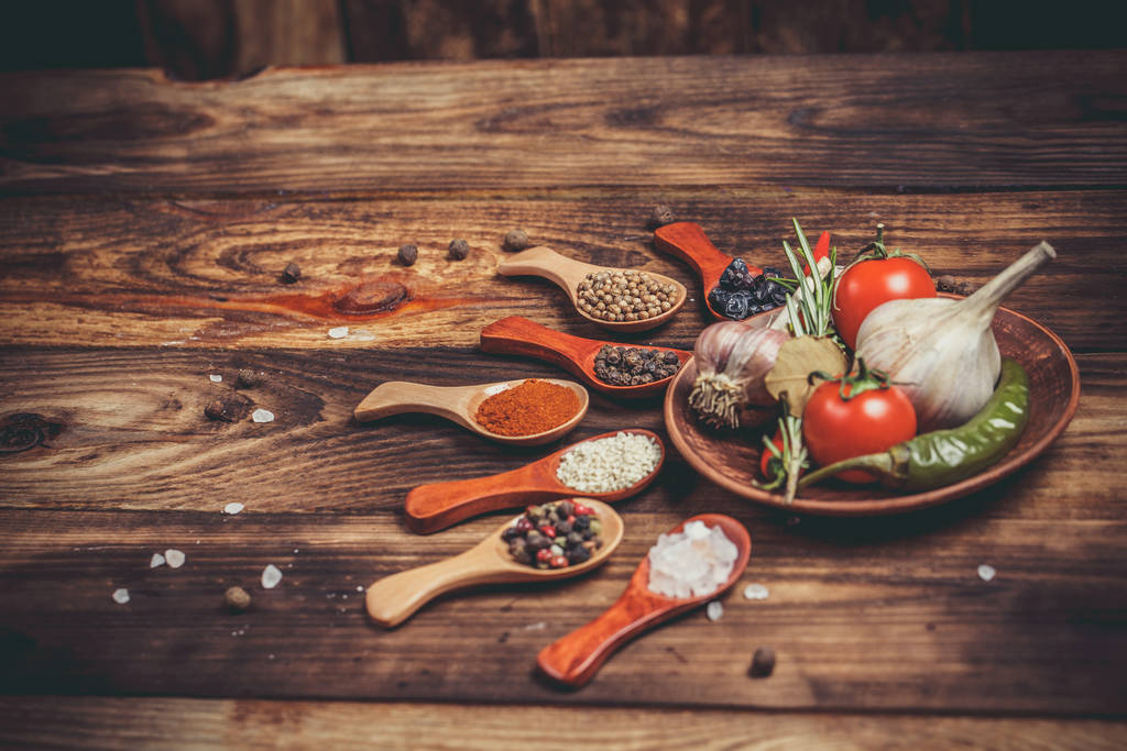 Verschillende specerijen in houten lepels en verschillende ingrediënten voor koken, kerstomaten, knoflook, koriander en rode peper - Foto, afbeelding