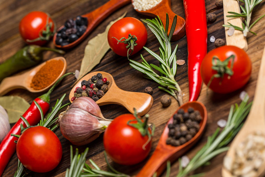 Verschillende specerijen in houten lepels en verschillende ingrediënten voor koken, kerstomaten, knoflook, koriander en rode peper - Foto, afbeelding