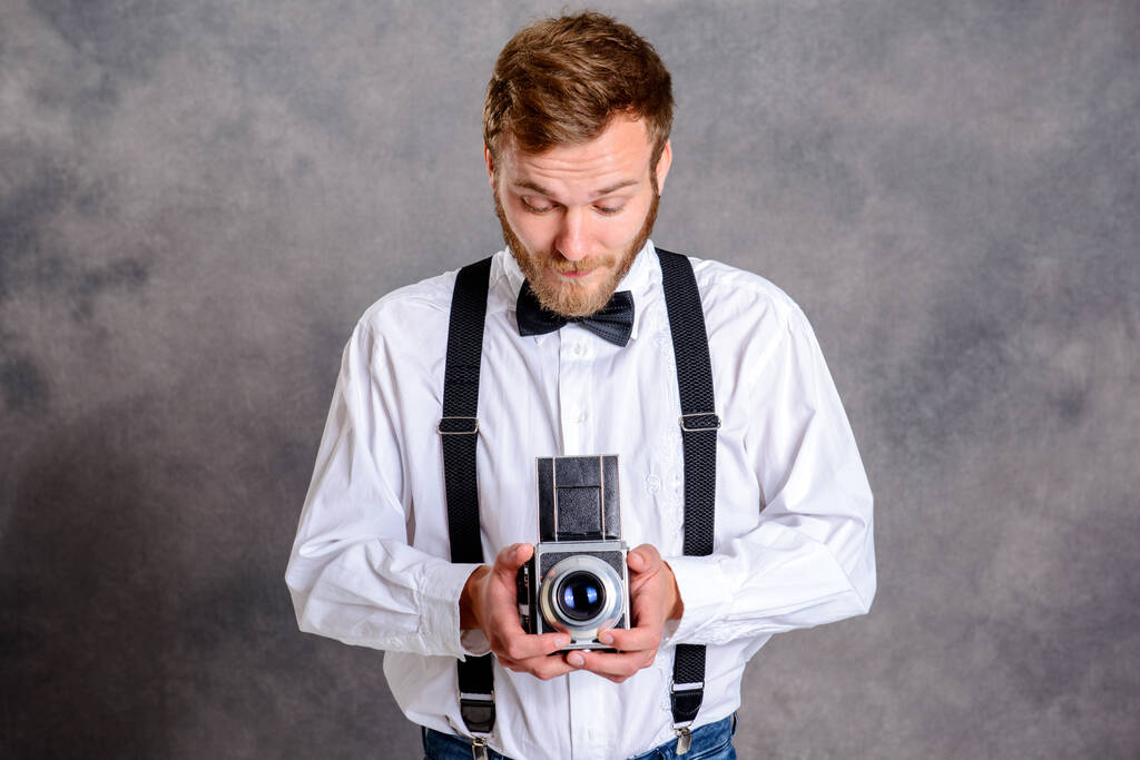 joven barbudo con camisa blanca y pajarita con cámara vieja delante de fondo gris
 - Foto, Imagen