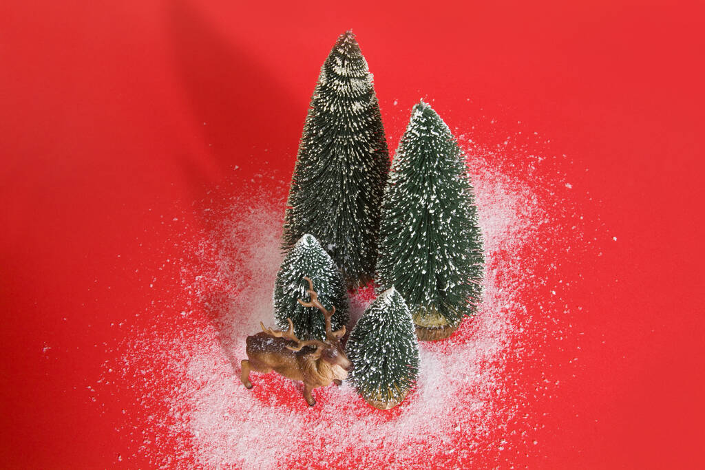 Μεμονωμένη ομάδα από πλήρως τεχνητά έλατα σαν ένα μικρό χιονισμένο δασικό δέντρο με ένα αγαλματίδιο ταράνδων μέσα σε ένα ζωηρό κόκκινο φόντο. Ελάχιστη φωτογραφία νεκρής φύσης - Φωτογραφία, εικόνα