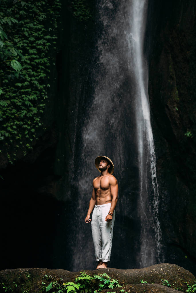 Мужчина спортивного телосложения у водопада. Человек путешествует по миру. Человек у водопада. Путешествие на Бали Индонезия. Мужчина в конической шляпе и белых штанах у водопада. Одинокий путешественник
. - Фото, изображение