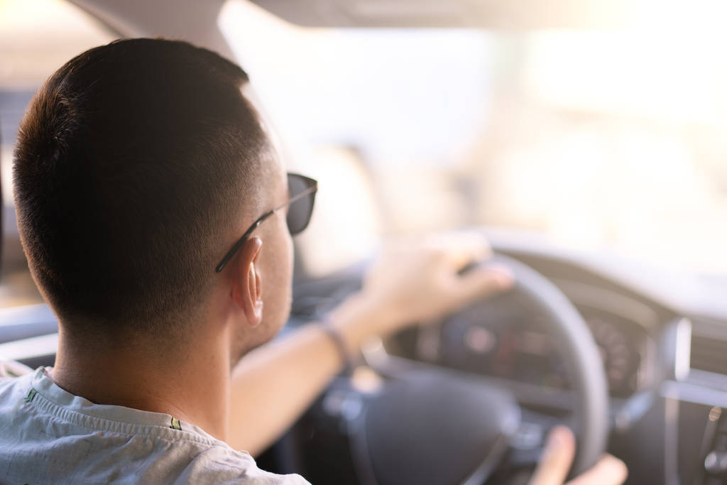 Νεαρός Καυκάσιος άνδρας κάθεται σε ένα αυτοκίνητο πίσω όψη κρατώντας το τιμόνι; βλέποντας τη μετάφραση σε μια φωτεινή ηλιόλουστη μέρα - Φωτογραφία, εικόνα