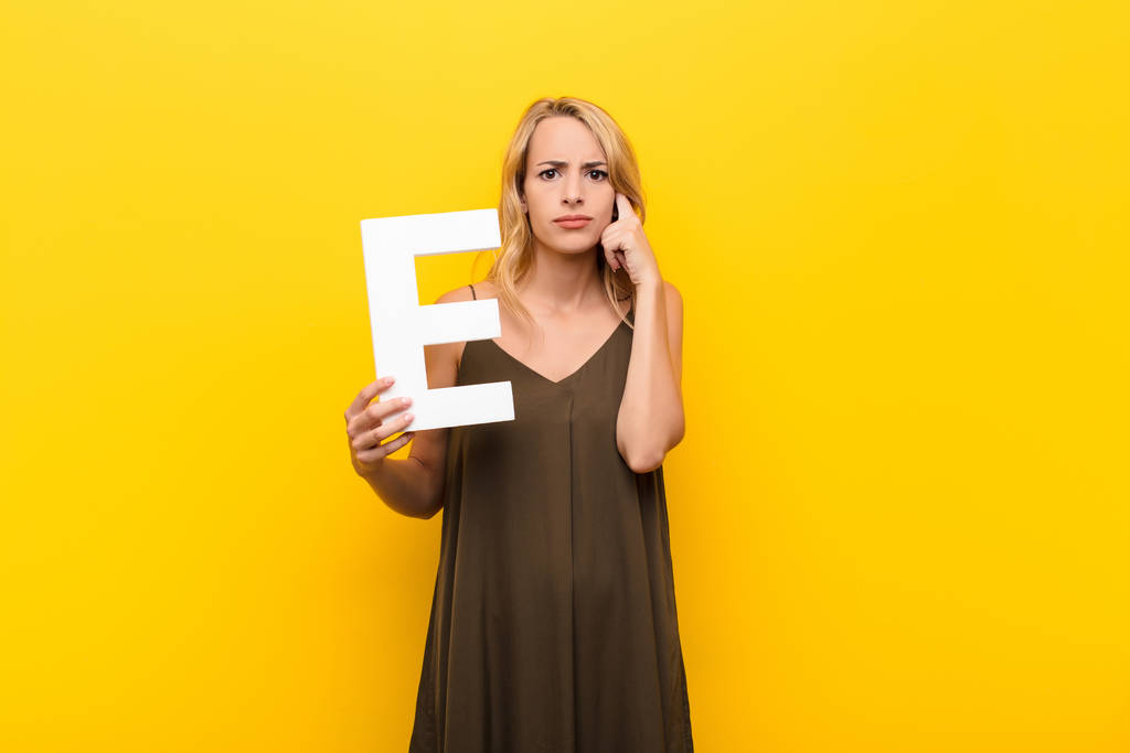 młoda blondynka zdezorientowana, wątpliwa, myśląca, trzymająca literę E alfabetu, aby utworzyć słowo lub zdanie. - Zdjęcie, obraz