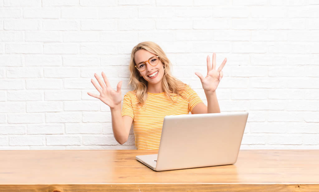 junge blonde Frau lächelt und sieht freundlich aus, zeigt die Zahl neun oder neun mit der Hand nach vorne und zählt mit einem Laptop herunter - Foto, Bild