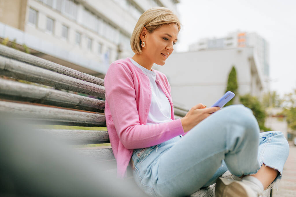 Πλάγια άποψη εικόνα της όμορφης ξανθιάς νεαρής γυναίκας που φοράει μπλε τζιν, λευκό t-shirt και ροζ σακάκι, χαμογελώντας και στέλνοντας μηνύματα στο έξυπνο τηλέφωνο κάθεται στον πάγκο στο δρόμο της πόλης την άνοιξη. - Φωτογραφία, εικόνα