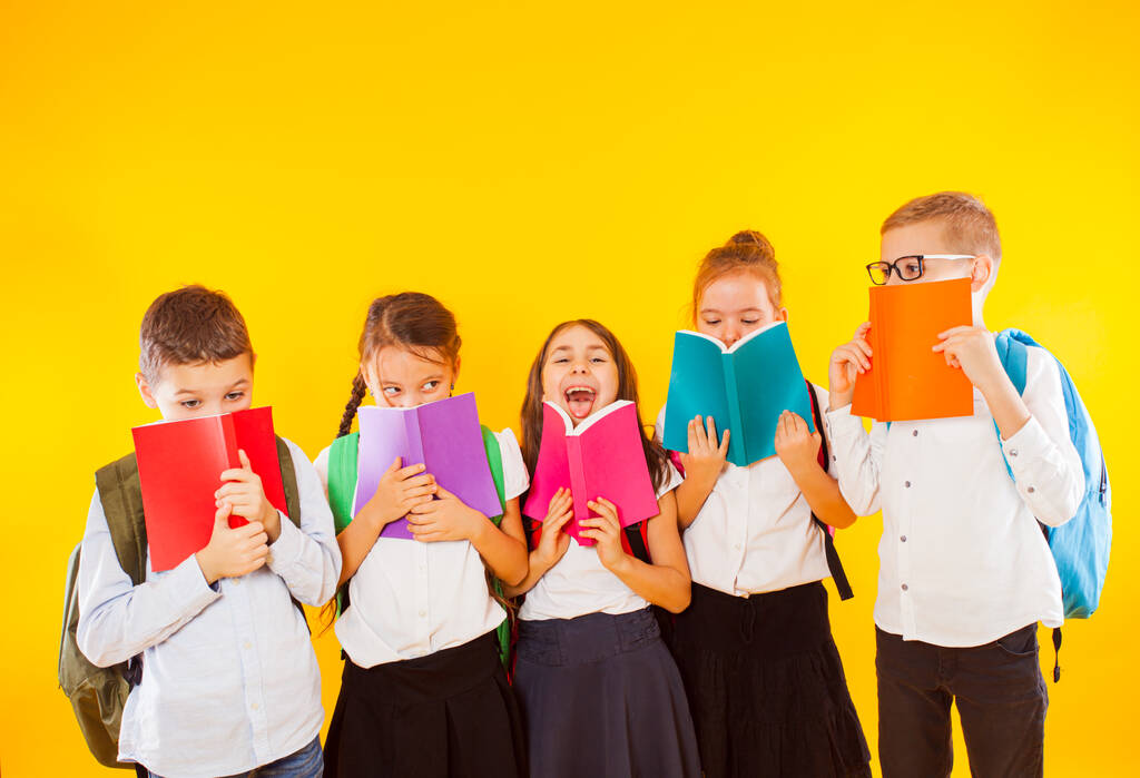 Ευτυχισμένα σχολιαρόπαιδα στέκονται με πολύχρωμα βιβλία απομονωμένα σε κίτρινο φόντο. Τα παιδιά κρύβονται πίσω από βιβλία. - Φωτογραφία, εικόνα