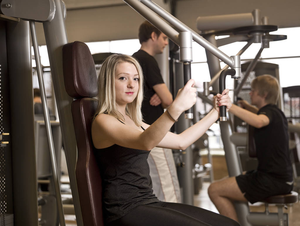 Chica usando una máquina de ejercicios en un club de salud con dos hombres en el fondo
 - Foto, imagen