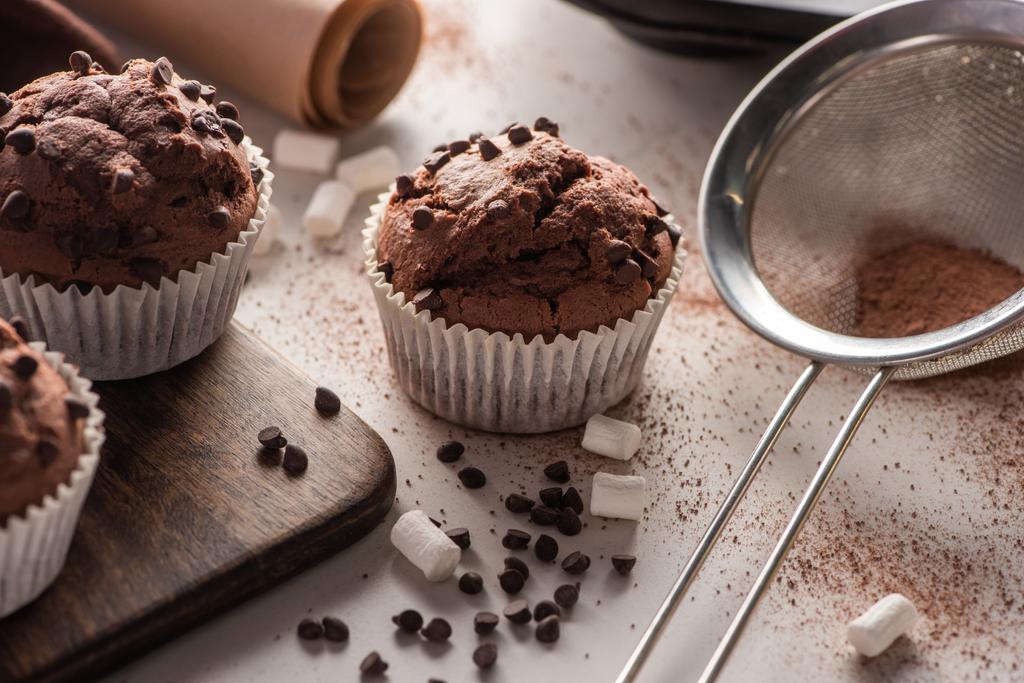 close up view of fresh chocolate muffins near marshmallow, cocoa powder and sieve on wooden cutting board - Photo, Image