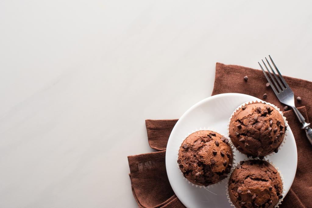 vrchní pohled na čerstvé čokoládové muffiny na bílém talíři a hnědý ubrousek u vidličky na mramorovém povrchu - Fotografie, Obrázek
