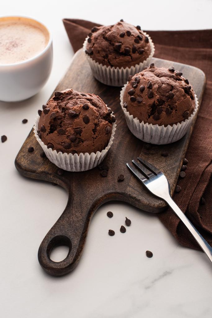 fresh chocolate muffins on wooden cutting board near napkin, fork and coffee - Fotoğraf, Görsel