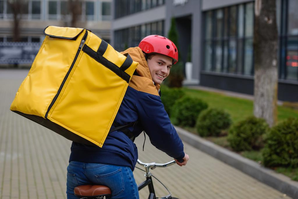 Rückansicht des Lieferanten mit Thermo-Rucksack, der auf der Stadtstraße mit dem Fahrrad in die Kamera lächelt - Foto, Bild
