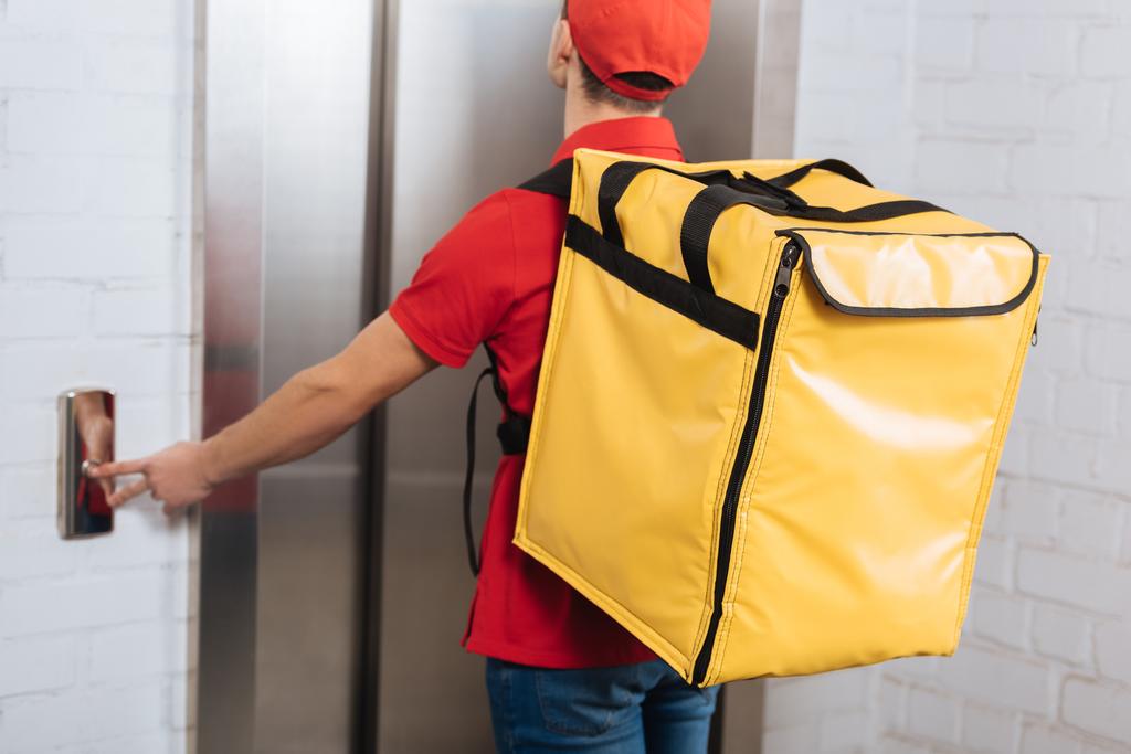 Vista trasera del repartidor hombre en uniforme rojo con la mochila térmica presionando el botón del ascensor
  - Foto, Imagen