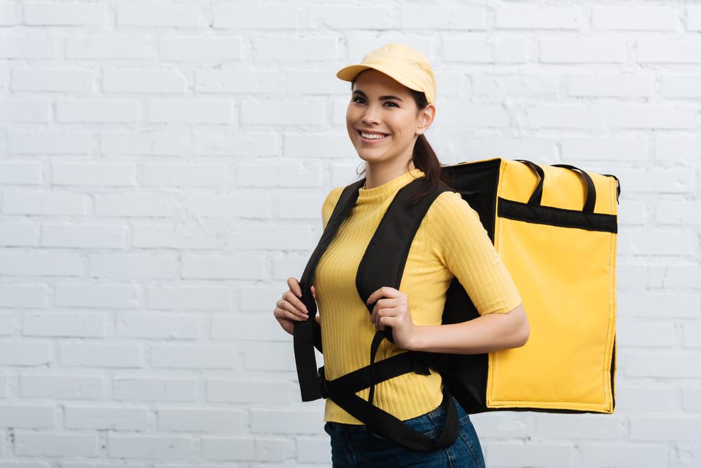 Belo mensageiro em uniforme amarelo carregando mochila térmica e sorrindo para a câmera perto da parede de tijolo
 - Foto, Imagem