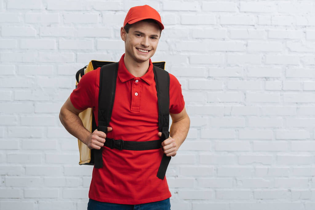 Zusteller in roter Uniform mit Thermo-Rucksack und lächelnd in die Kamera nahe Ziegelmauer - Foto, Bild