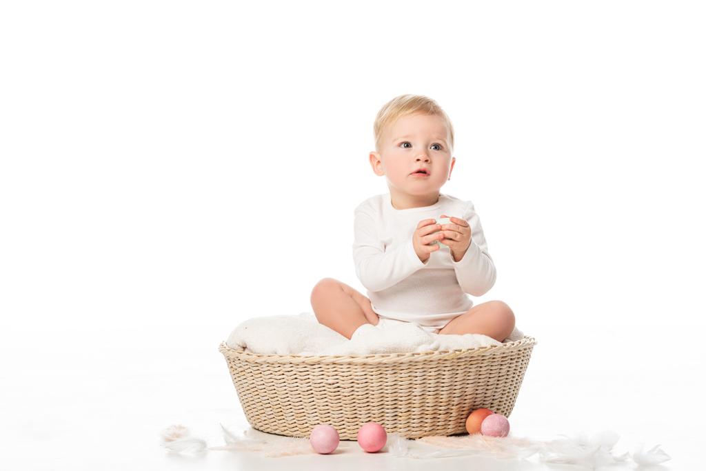 Criança segurando ovo de Páscoa, sentado em cesta com decoração colorida em torno de fundo branco
 - Foto, Imagem