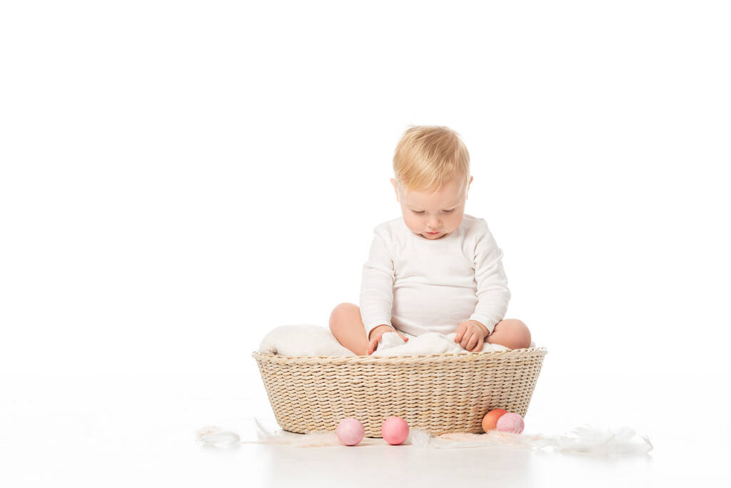 Child with lowered head on blanket in basket with Easter eggs around on white background - Photo, Image