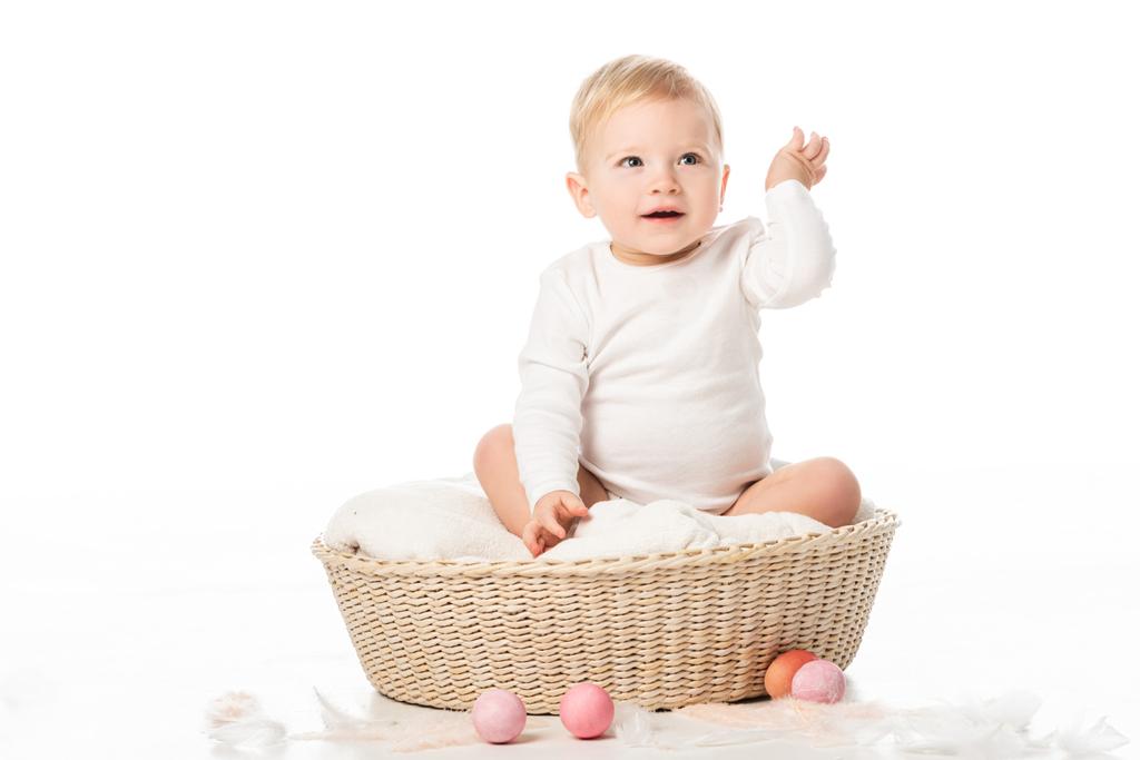 Criança com a mão levantada sentada no cobertor na cesta com ovos de Páscoa ao redor no fundo branco
 - Foto, Imagem