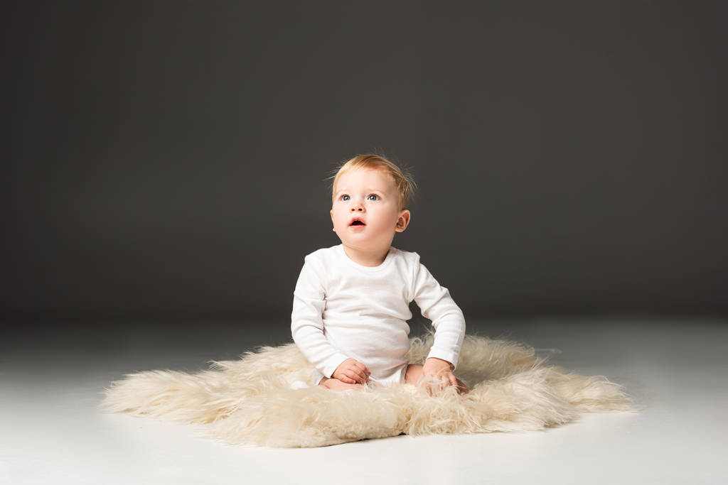 Cute kid looking up with open mouth, sitting on fur on black background - Photo, Image