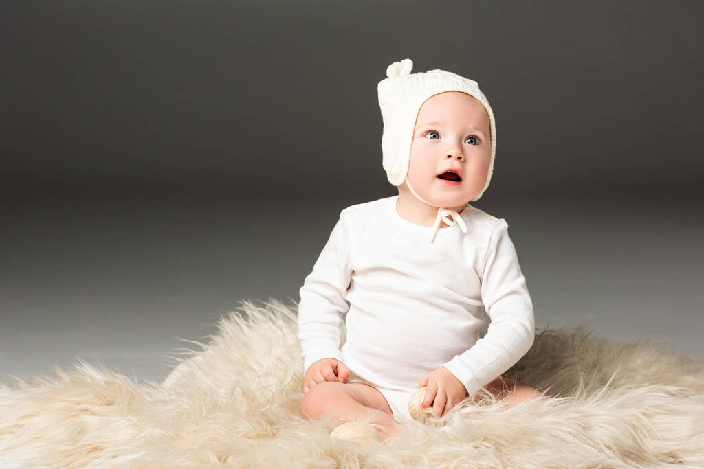 Child wearing baby bonnet, holding easter egg with open mouth on fur on black background - Photo, Image