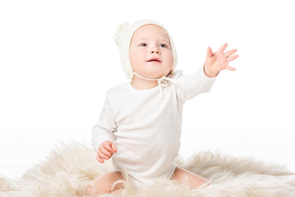 Παιδί που φοράει μωρουδίστικο καπέλο, κοιτάζει ψηλά με απλωμένο χέρι, κάθεται πάνω σε γούνα απομονωμένο πάνω σε λευκό  - Φωτογραφία, εικόνα