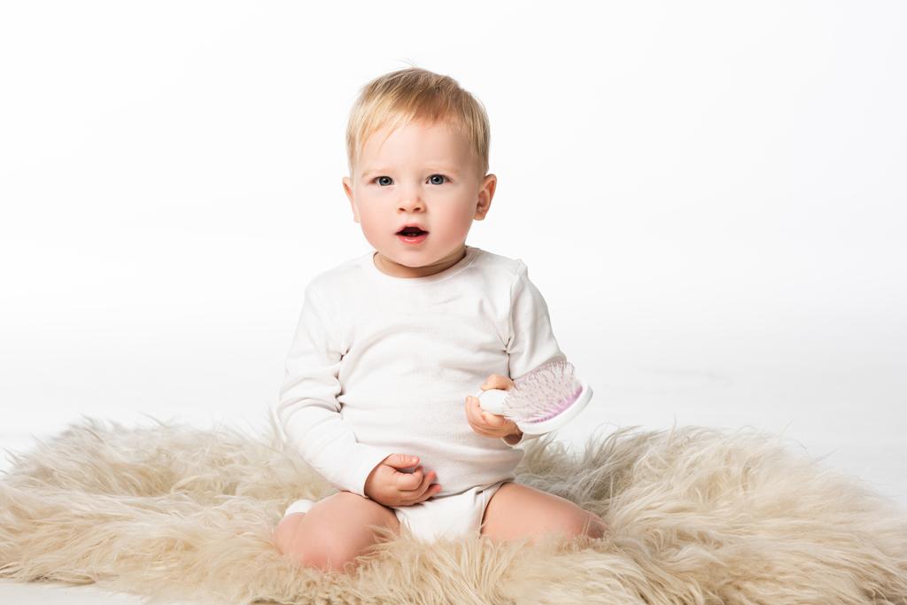 Kid looking at camera with open mouth holding comb and sitting on fur isolated on white  - Photo, Image