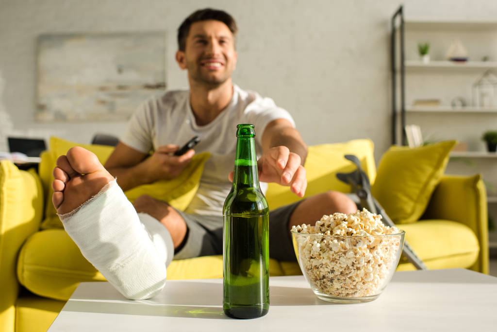 Selektiver Fokus eines lächelnden Mannes mit gebrochenem Bein, der auf der Couch neben Popcorn Bier trinkt - Foto, Bild