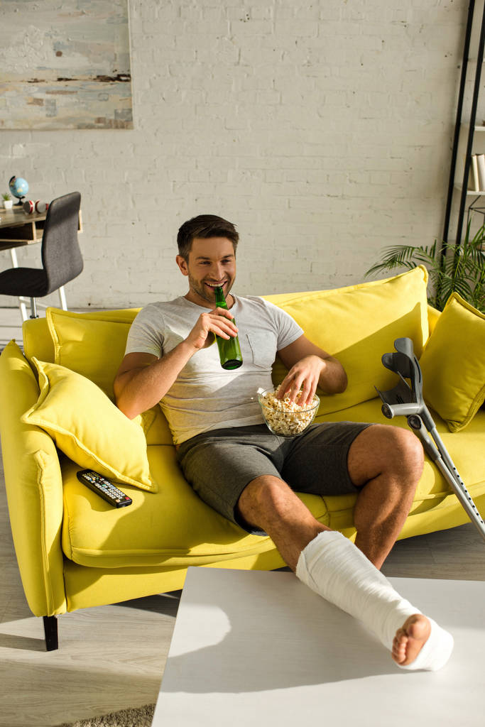 Sonriente hombre con la pierna rota bebiendo cerveza y sosteniendo palomitas de maíz mientras ve la película en el sofá
 - Foto, Imagen